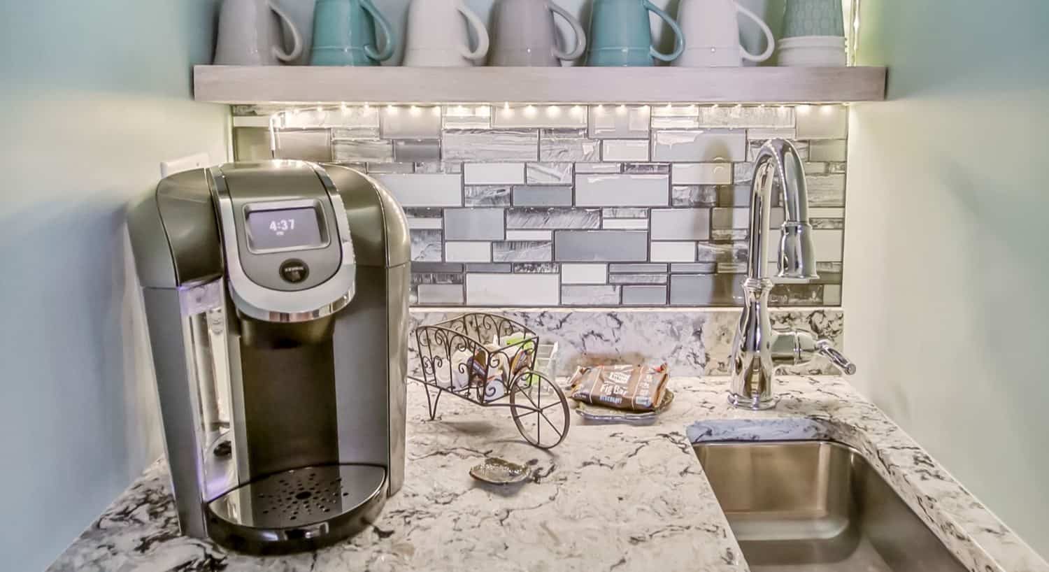 Marble counter top with tiled backsplash, coffee maker, small sink, and gray, white, and teal coffee cups