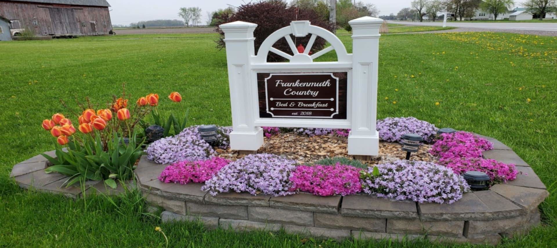 Welcome sign for the property surrounded by orange, purple, and pink flowers in raised stone garden bed surrounded by green grass