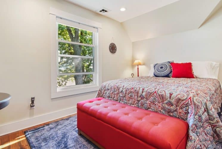 Bedroom with light cream walls, hardwood flooring, bed with navy, red, and tan paisley bedding, and large red cushioned storage box at the foot of the bed